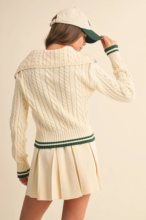 Woman wearing a cream cable knit sweater and skirt with a green and white cap on a beige background