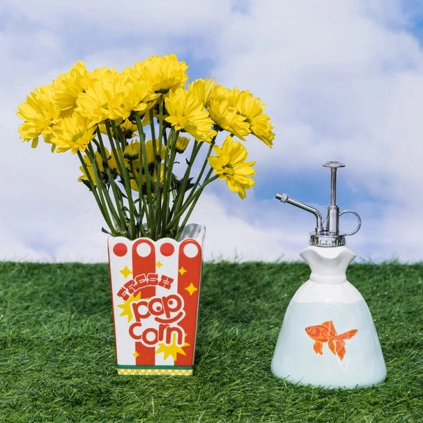 Yellow flowers in a popcorn box with a white sprayer on grass against a blue sky.
