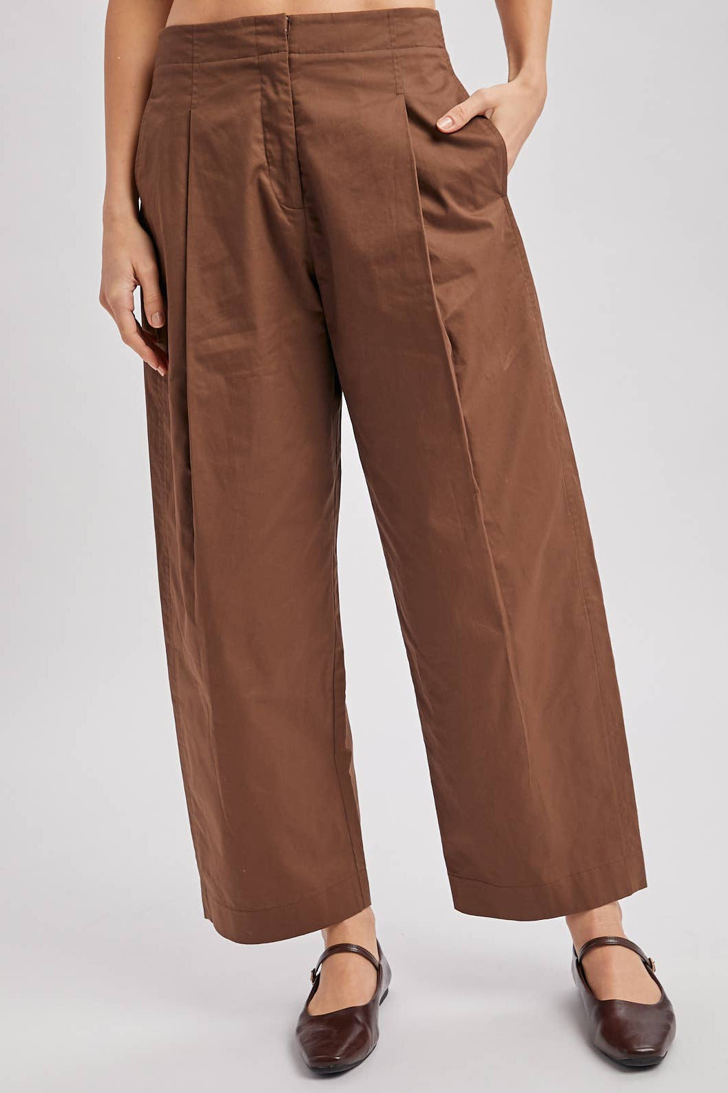A person wearing brown wide-leg cropped pants with front pleats and side pockets, paired with dark brown flat shoes, against a light gray studio background.