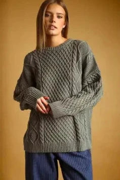 A woman wearing a chunky cable-knit sweater in a muted gray-green color, paired with blue striped pants, set against a solid warm brown background.