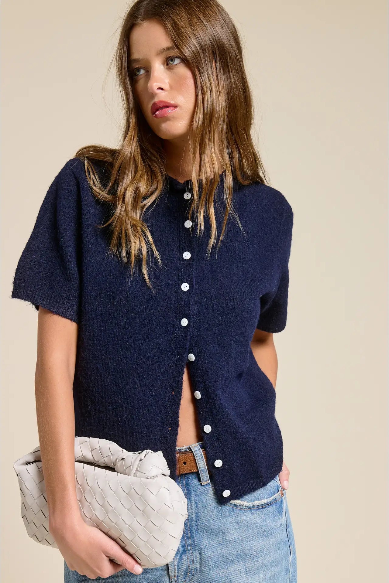 Woman wearing a navy blue cardigan with cut-out design, holding a beige clutch against a beige background.