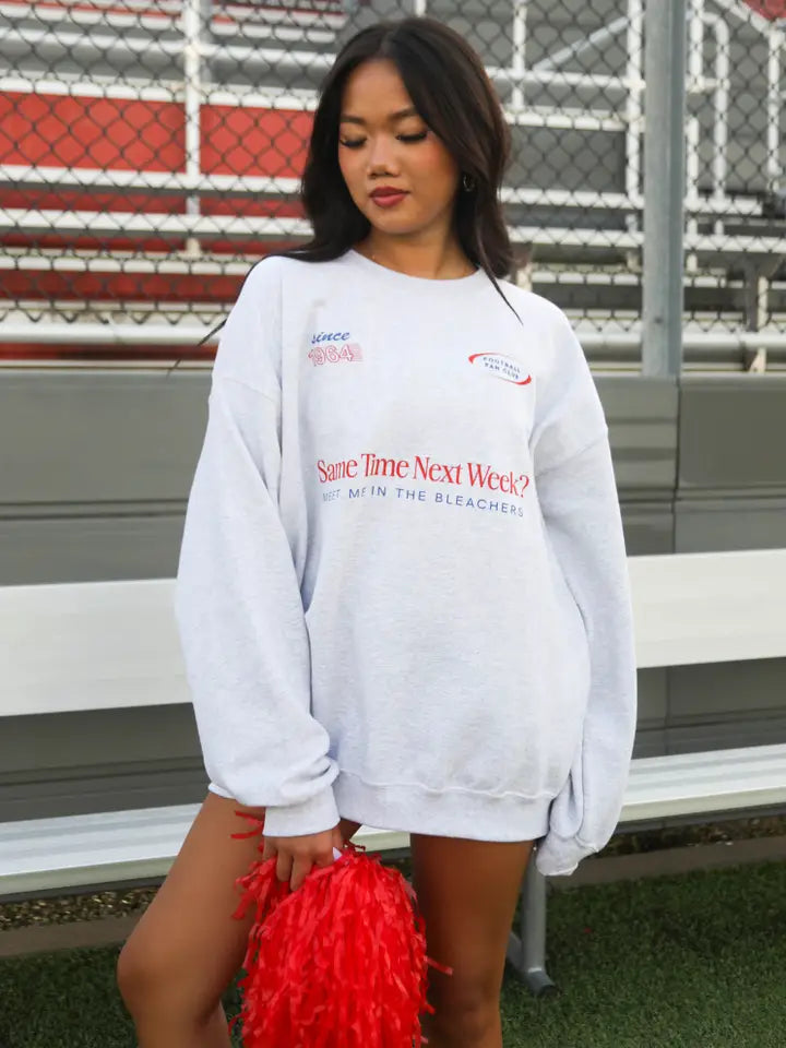 Woman wearing a white sweatshirt with text, holding a red pom-pom in front of bleachers.