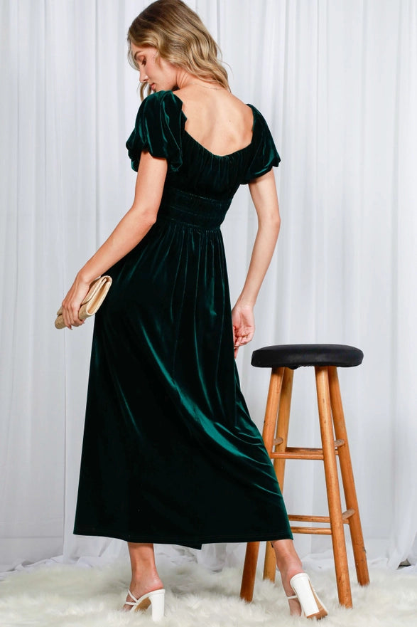 Woman wearing a green velvet dress standing next to a wooden stool against a white curtain background.