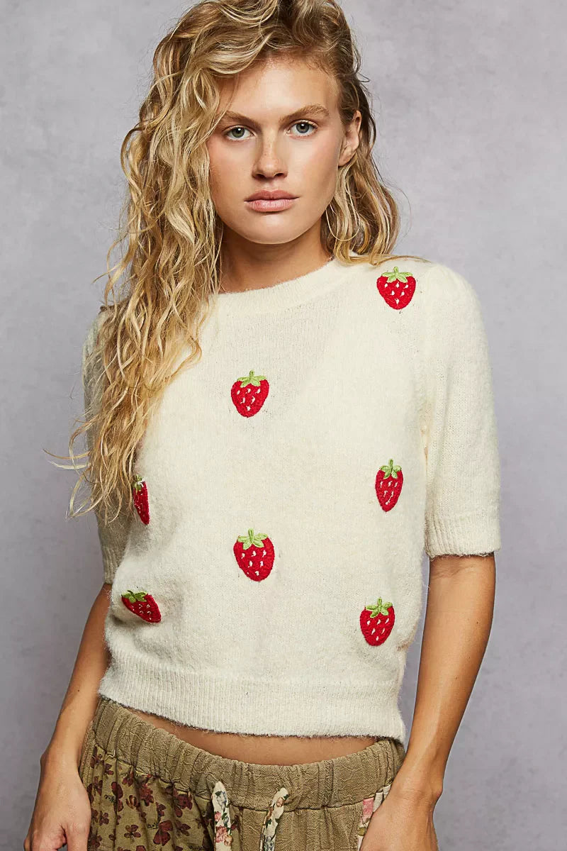 Woman wearing a cream sweater with strawberry patterns against a gray background