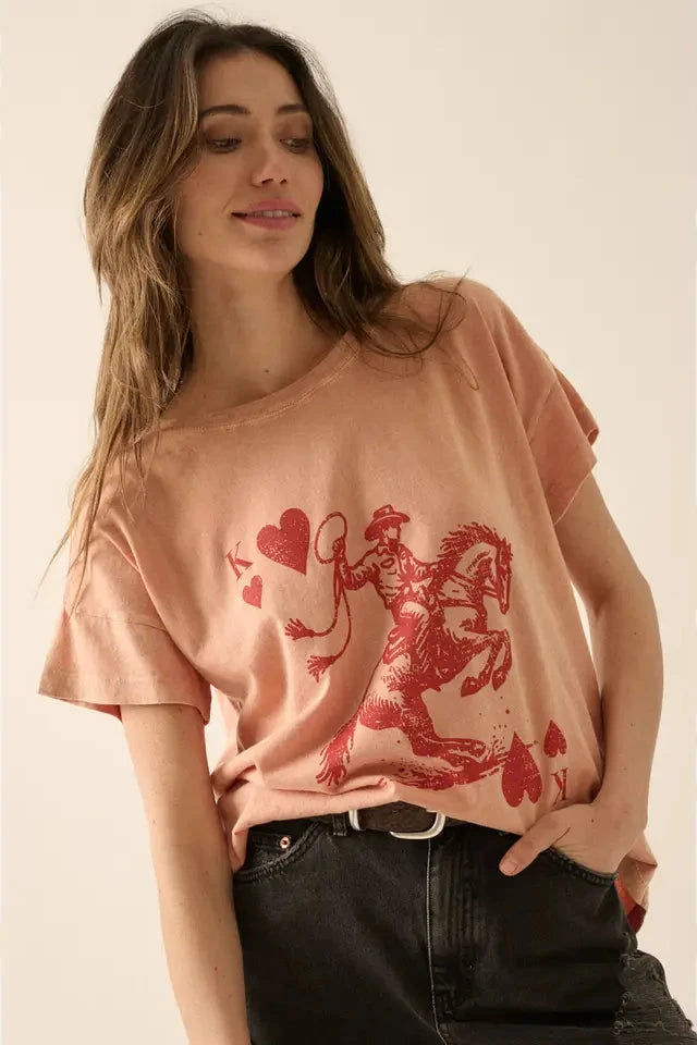 A woman wearing a pink graphic t-shirt with a vintage-style cowboy king of hearts playing card graphic, round neckline, short sleeves, and a relaxed fit.