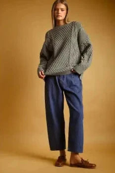 A model stands against a warm tan studio background wearing a chunky cable-knit sweater in a muted sage green, paired with dark blue wide-leg cropped trousers and brown leather loafers.