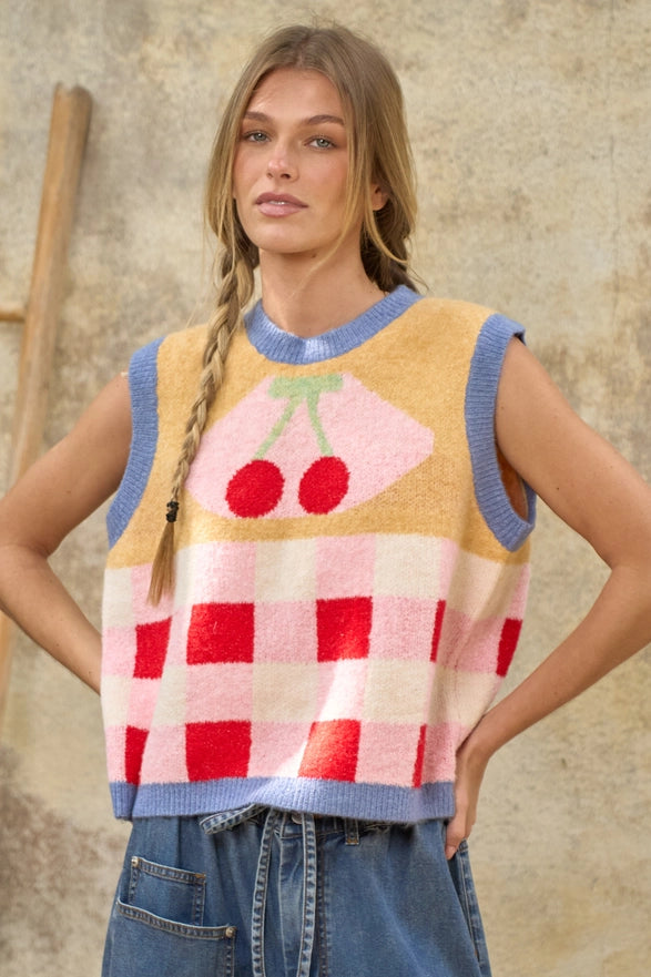 Woman wearing a colorful sleeveless sweater vest with cherry design against a textured wall.