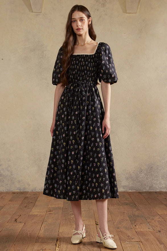 Woman wearing a black floral dress standing against a plain wall.