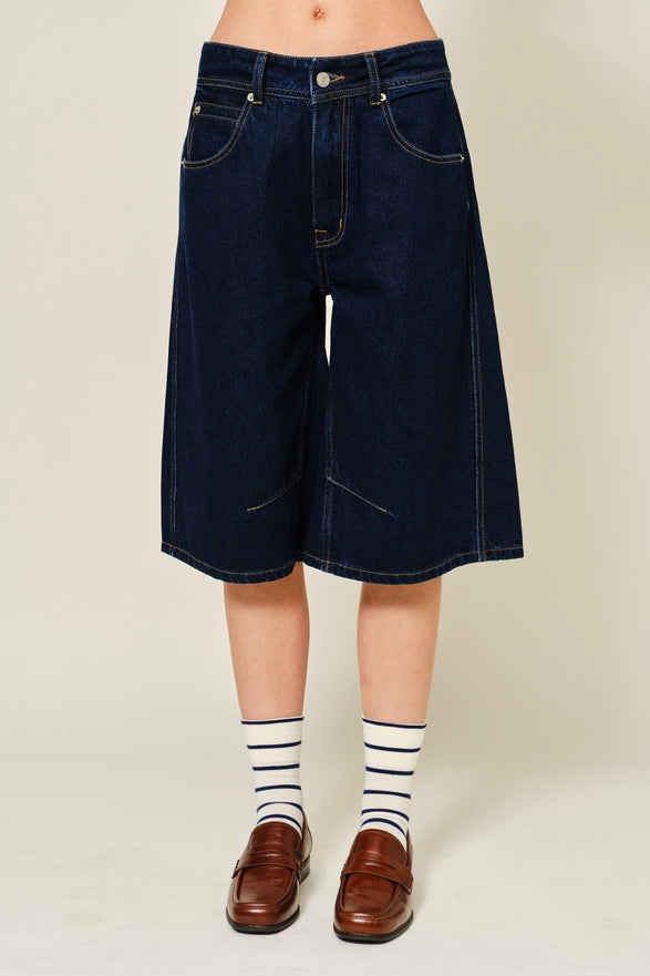 Person wearing dark blue denim shorts with striped socks and brown shoes on a plain background