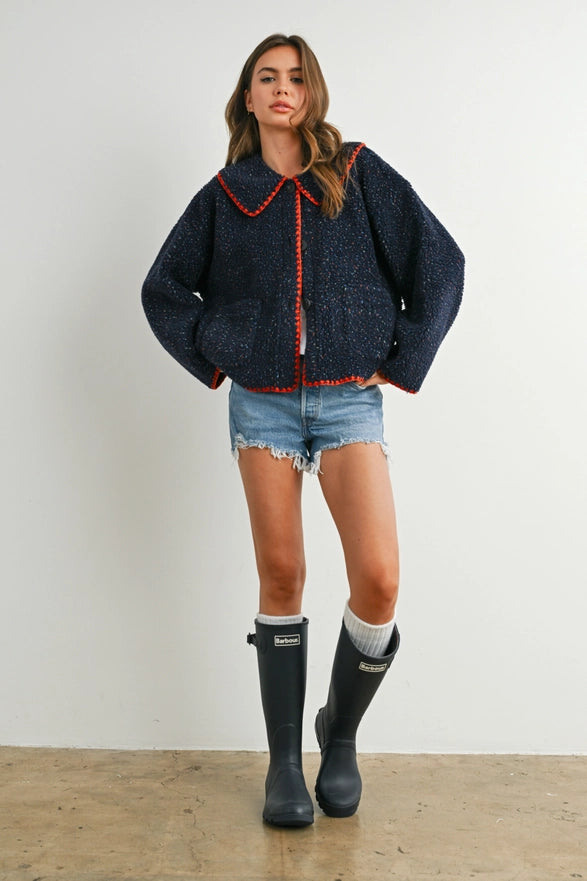 Woman wearing a dark blue jacket with red trim, denim shorts, and black rain boots on a white background.