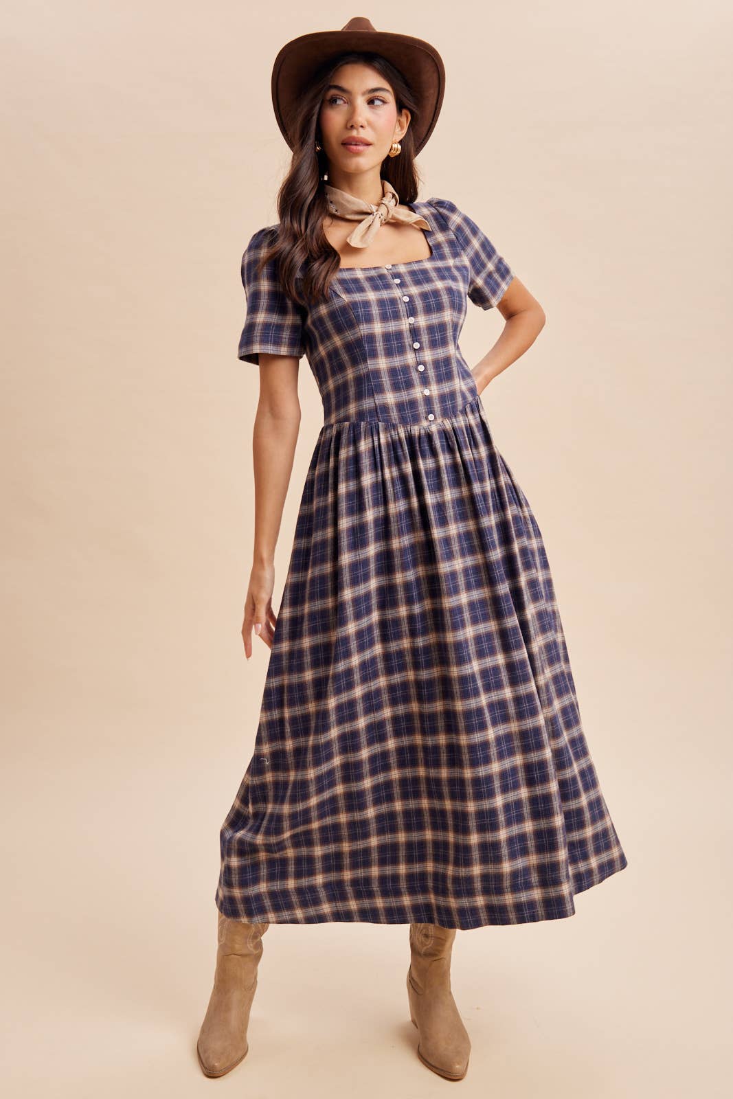 A woman modeling a short-sleeve plaid midi dress with a square neckline and button front, styled with a brown hat, beige scarf, and tan boots against a solid beige background.