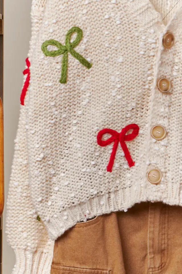 Close-up of a cream-colored knitted sweater with red and green bows on a plain background.