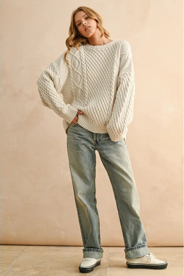 Woman wearing a white cable knit sweater and light blue jeans against a beige background