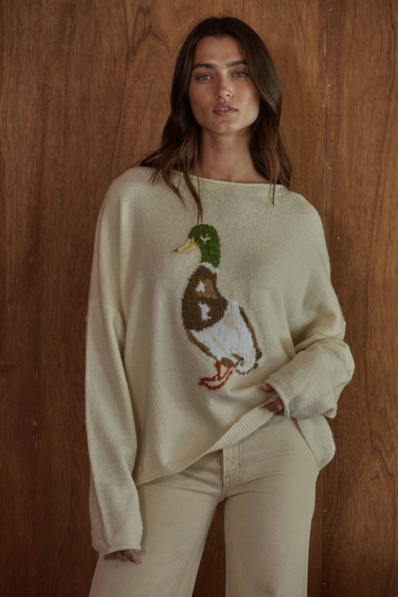 A woman wearing a cream-colored knit sweater with a large duck motif on the front, paired with matching cream pants, standing against a rustic wooden wall.