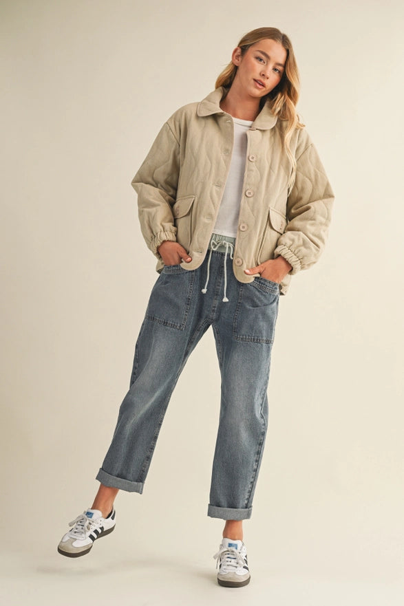 Woman wearing a beige jacket, white shirt, and blue jeans on a plain background