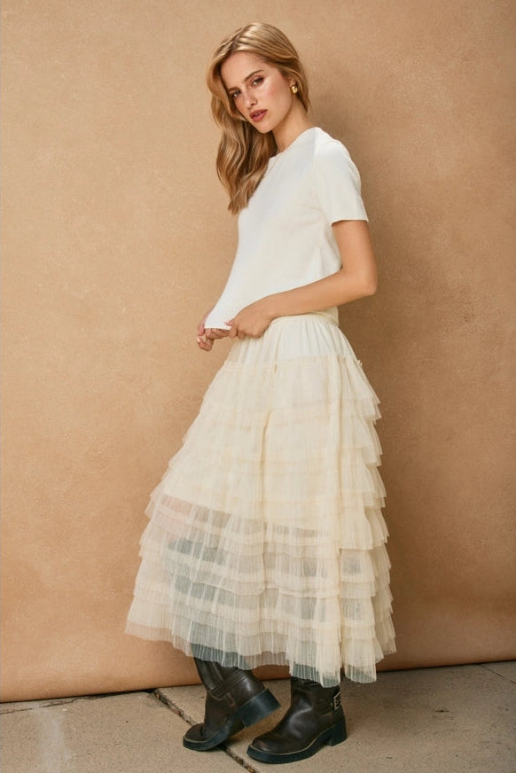 Woman wearing a white top and layered cream skirt against a beige wall.