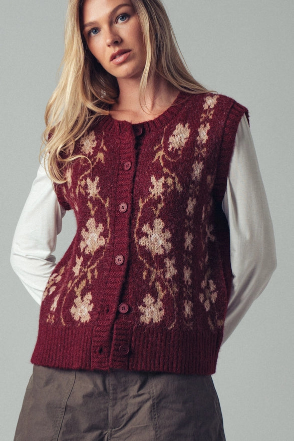 Woman wearing a patterned maroon vest over a white shirt on a gray background