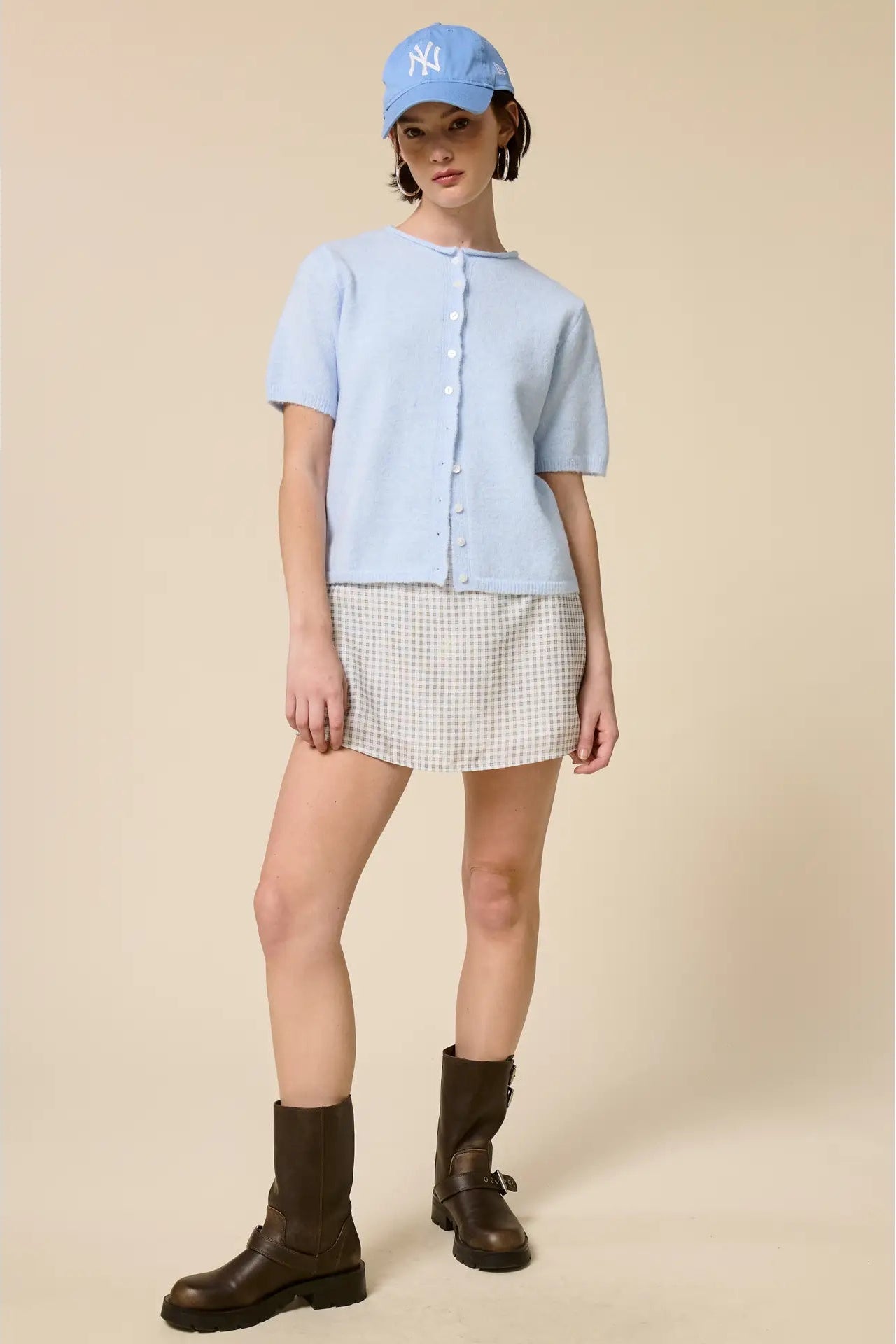 Person wearing a light blue shirt, checkered skirt, and brown boots on a beige background
