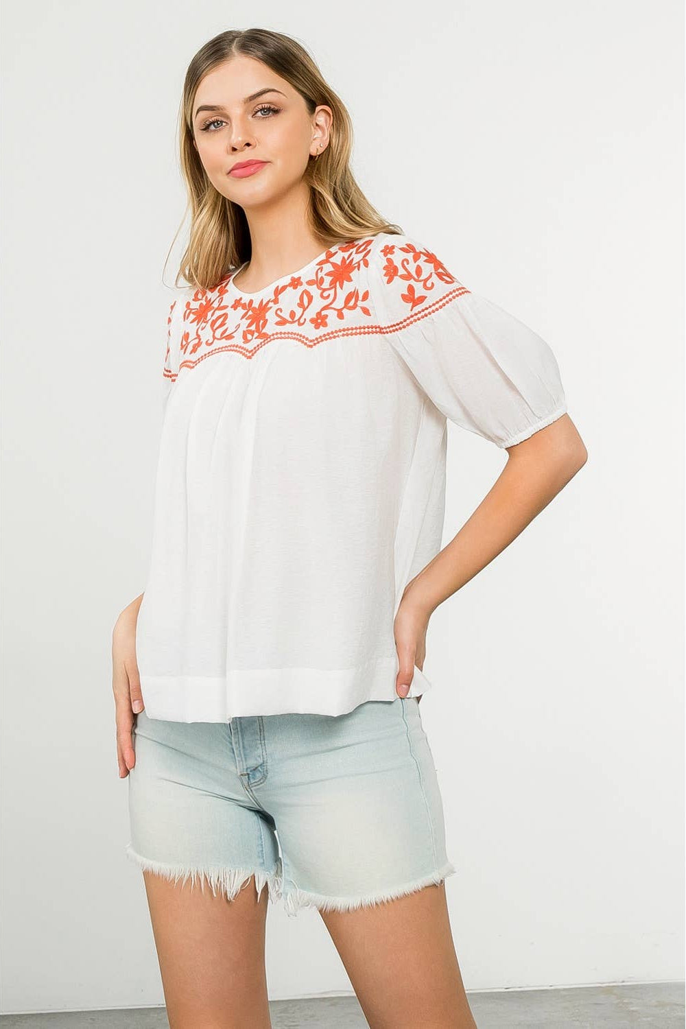 Woman wearing a white blouse with orange embroidery and denim shorts on a white background