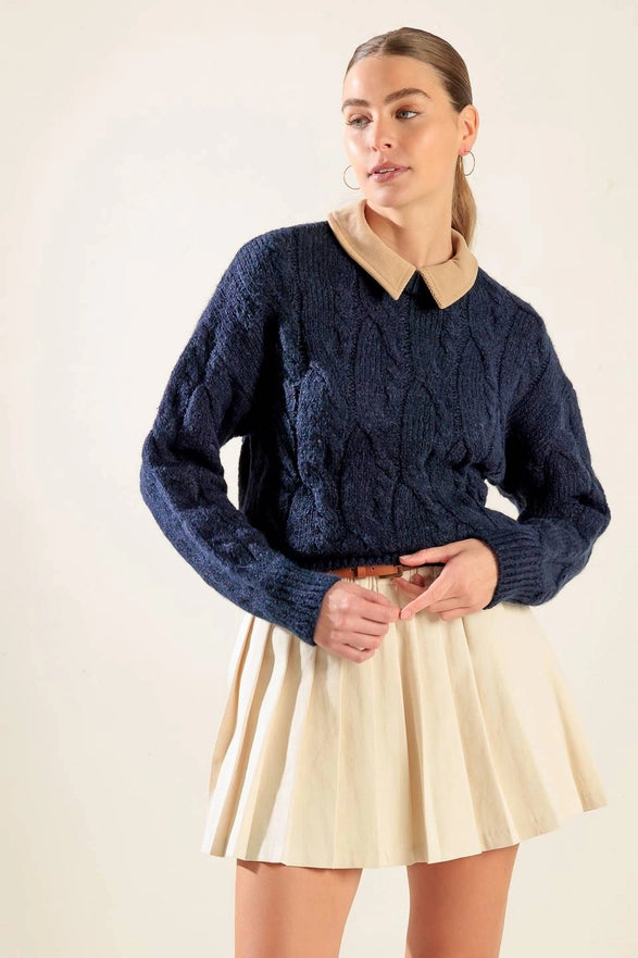 Woman wearing a navy blue cable knit sweater and beige pleated skirt on a plain background