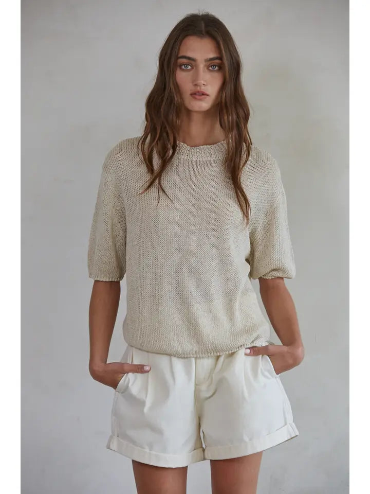 A woman wearing a beige short-sleeved sweater top with a round neckline, paired with white shorts.
