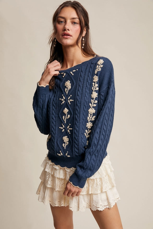 Woman wearing a blue sweater with floral embroidery and a beige skirt on a neutral background