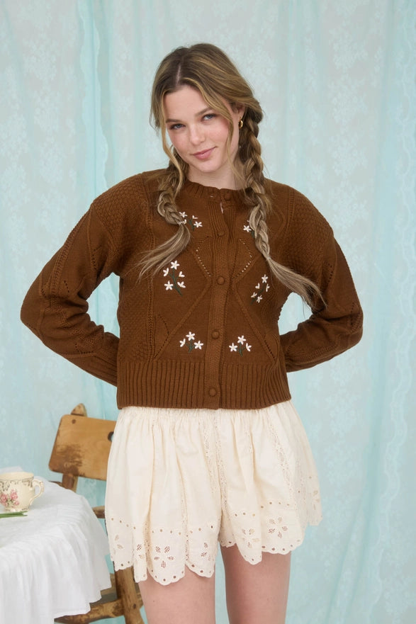 Woman wearing a brown cardigan with white patterns against a light blue curtain background