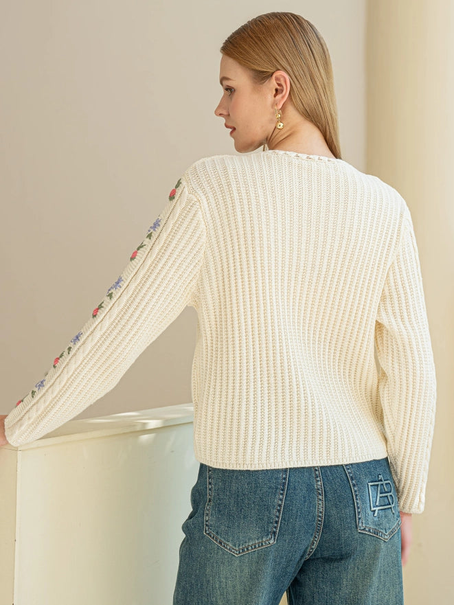 Woman wearing a cream ribbed sweater with floral sleeves and blue jeans against a neutral background