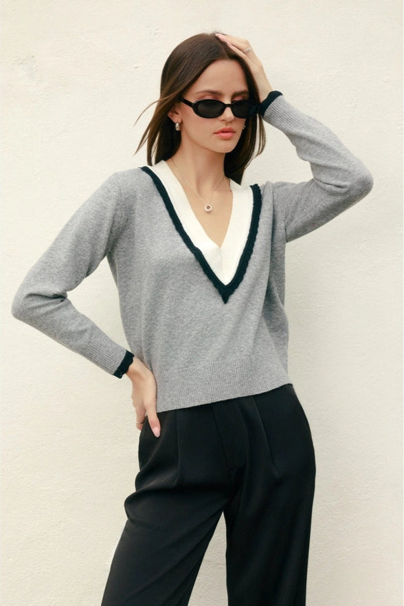 Woman wearing a gray sweater with a white collar and black trim, paired with black pants against a neutral background.