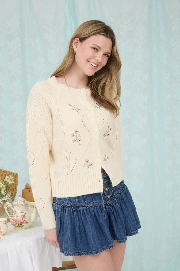 Woman wearing a cream sweater with floral embroidery and a denim skirt against a light blue curtain background.