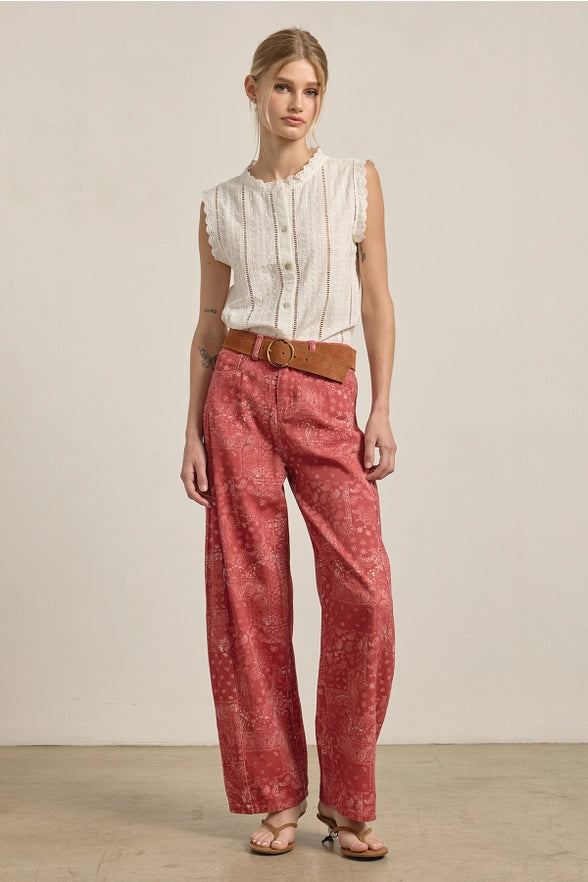 Woman wearing a white sleeveless blouse and red patterned pants with a brown belt against a plain background