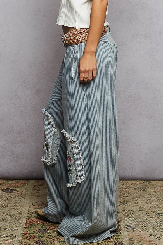 Person wearing blue striped pants with decorative pockets against a gray wall.