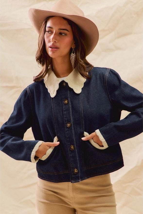 Woman wearing a navy denim jacket with white collar and cuffs, beige hat, and beige pants on a neutral background.