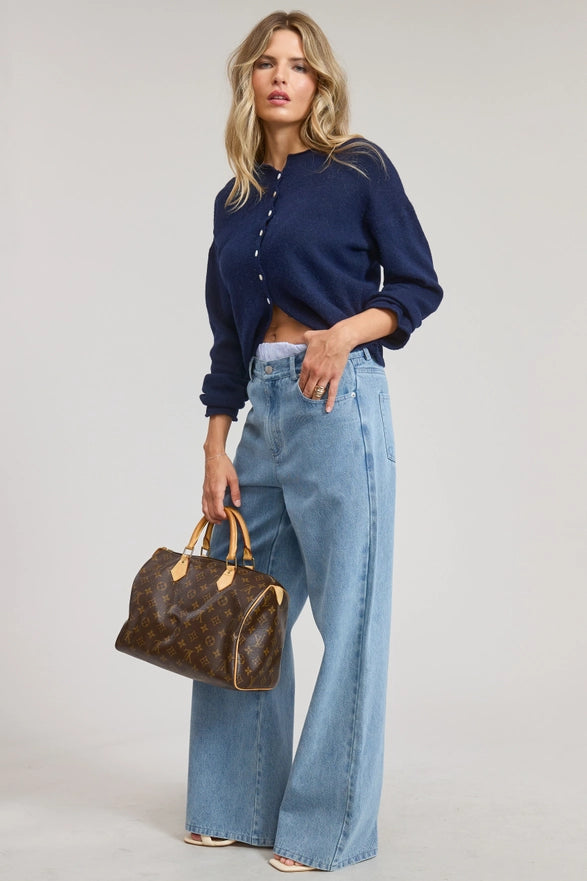 Woman wearing a navy shirt and blue jeans, holding a brown handbag on a light gray background
