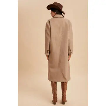 Person wearing a long beige coat and brown hat on a beige background