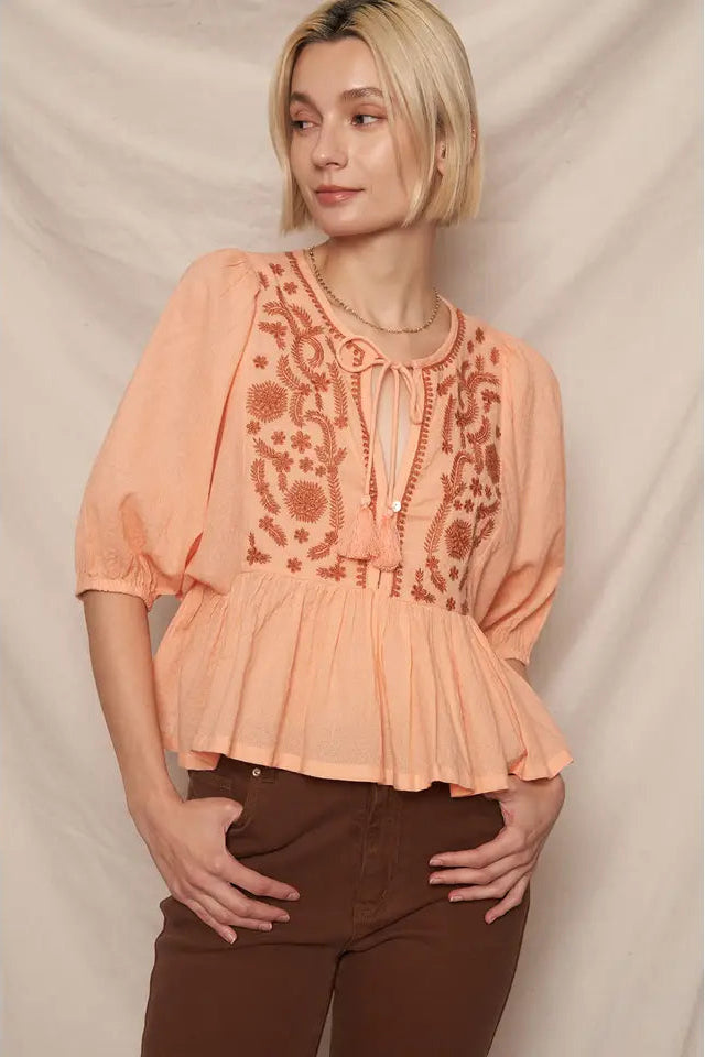 A woman wearing a peach-colored, puff sleeve peplum top with embroidery detailing, paired with brown pants.