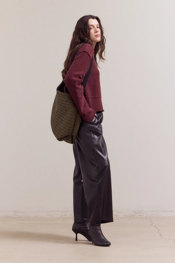 Woman wearing a maroon sweater, gray pants, and carrying a green bag against a plain background