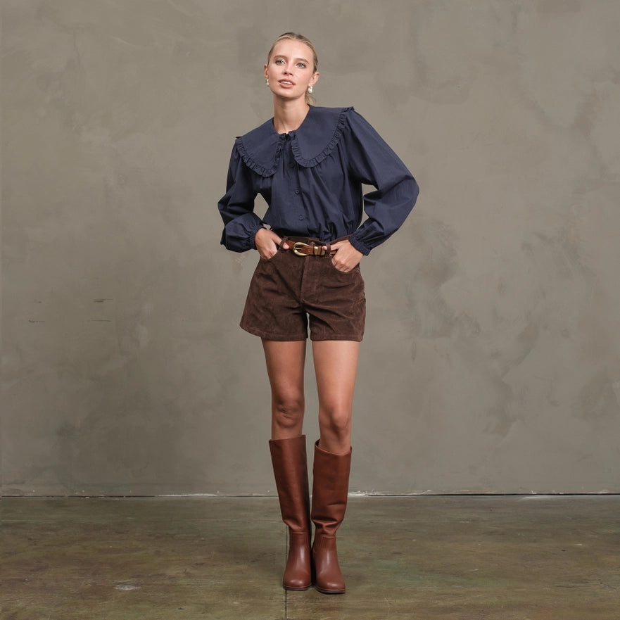 Woman wearing a dark blue blouse, brown shorts, and brown knee-high boots against a gray background