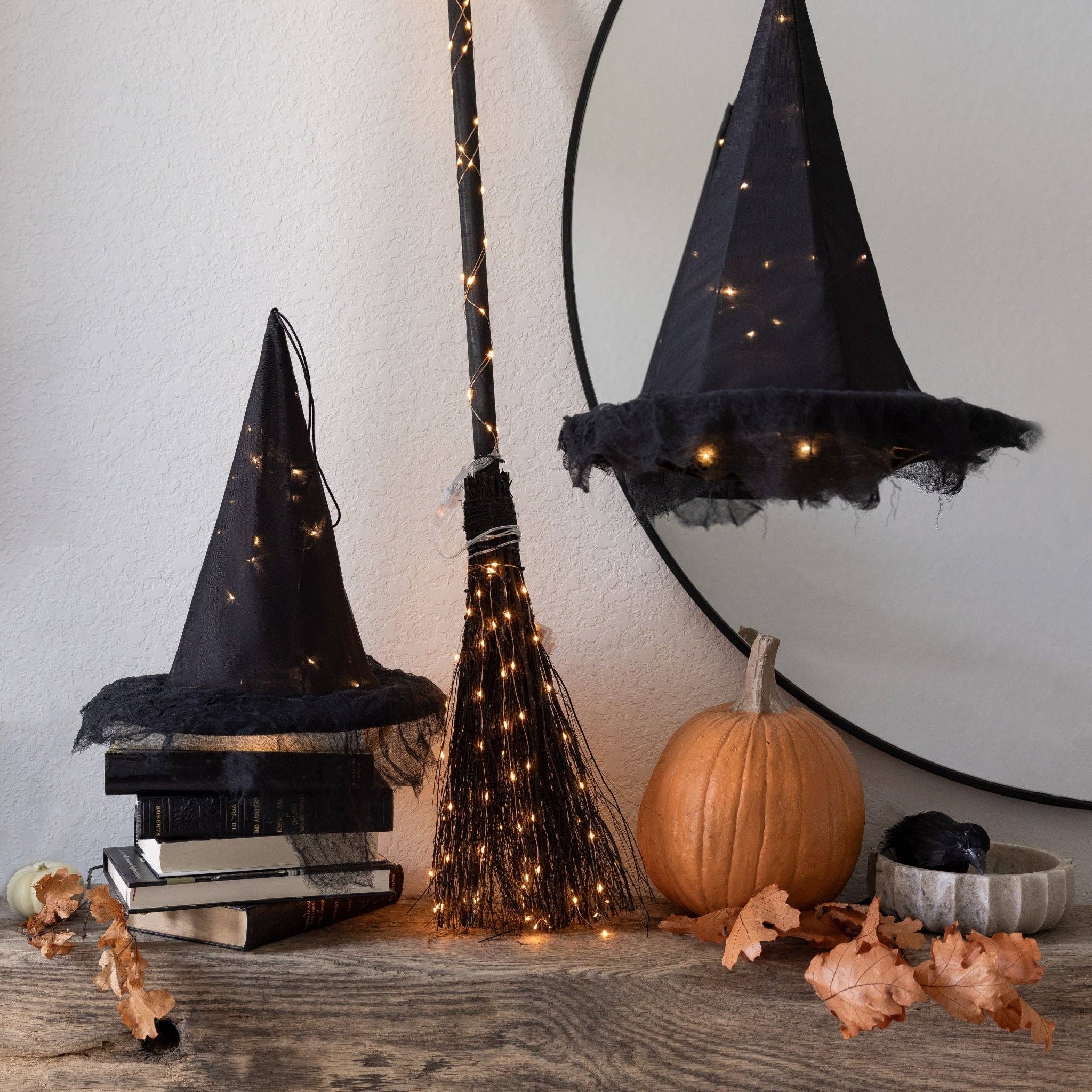 Halloween-themed black witch hat decorations with warm string lights, displayed on a rustic wooden surface alongside a broom, a small pumpkin, dried leaves, books, and a large round mirror reflecting the hats.