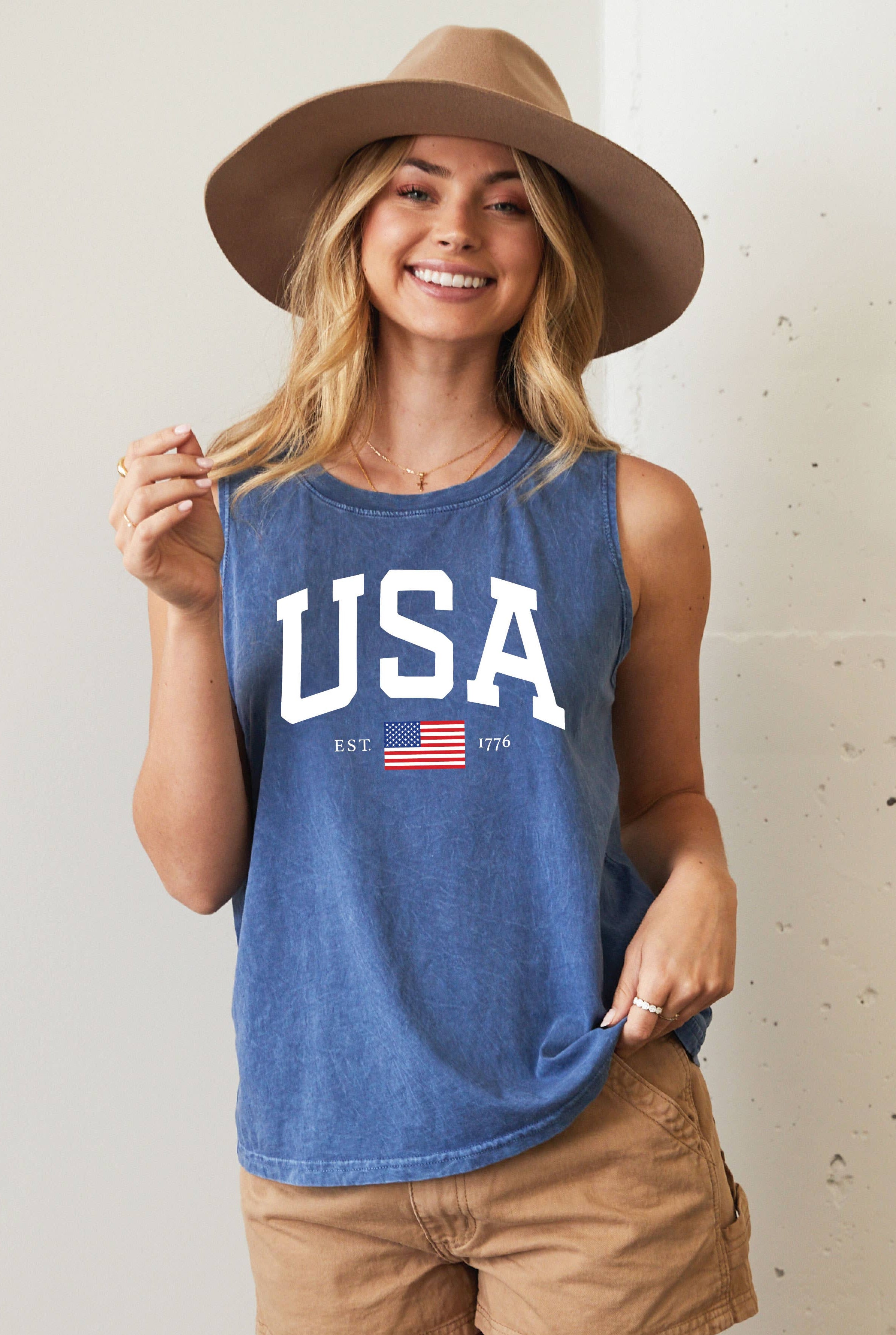 Woman wearing a blue 'SALTY' tank top and beige hat against a white wall.