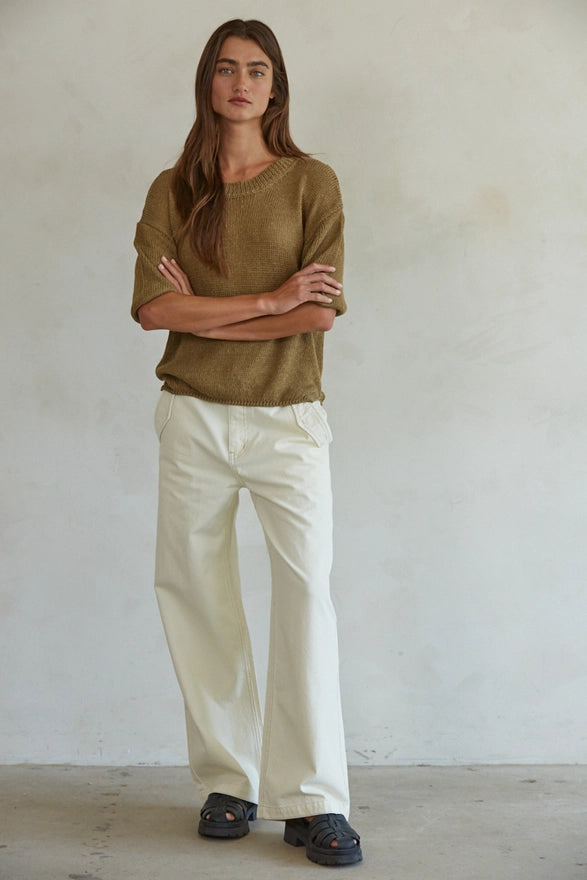 Person wearing a green sweater and white pants standing against a plain wall.