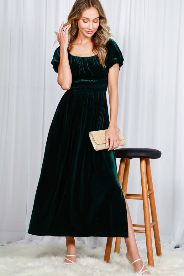 Woman wearing a green velvet dress holding a clutch against a white curtain background