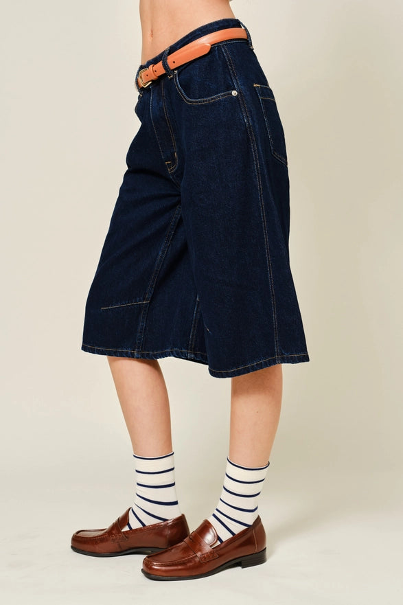Person wearing a dark denim skirt with a belt, brown shoes, and striped socks on a beige background