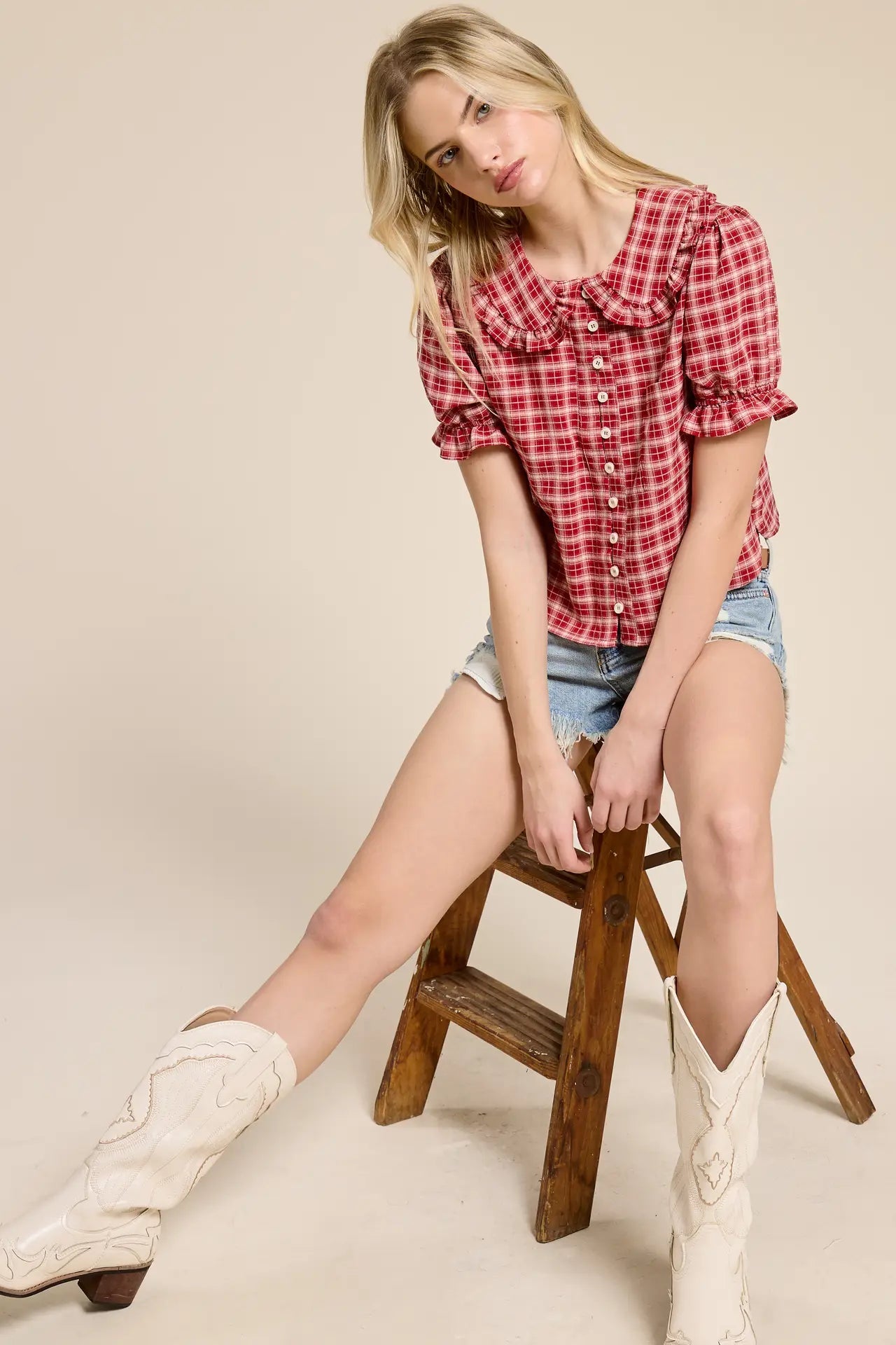 Woman wearing a red plaid shirt, denim shorts, and white cowboy boots sitting on a wooden stool.