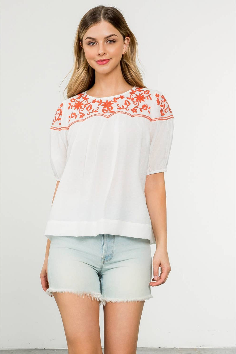 Woman wearing a white blouse with red embroidery and light blue shorts on a white background