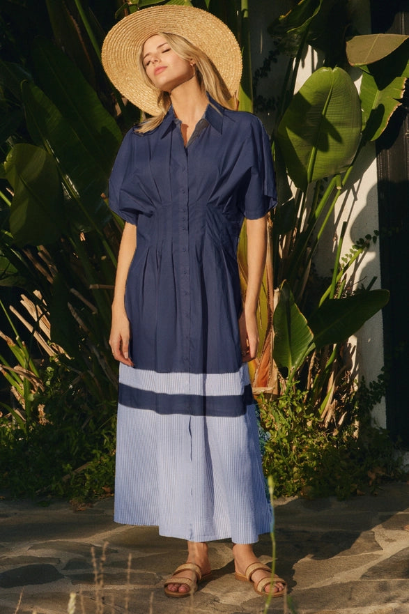 Woman wearing a navy and light blue dress with a straw hat, standing in a garden setting.