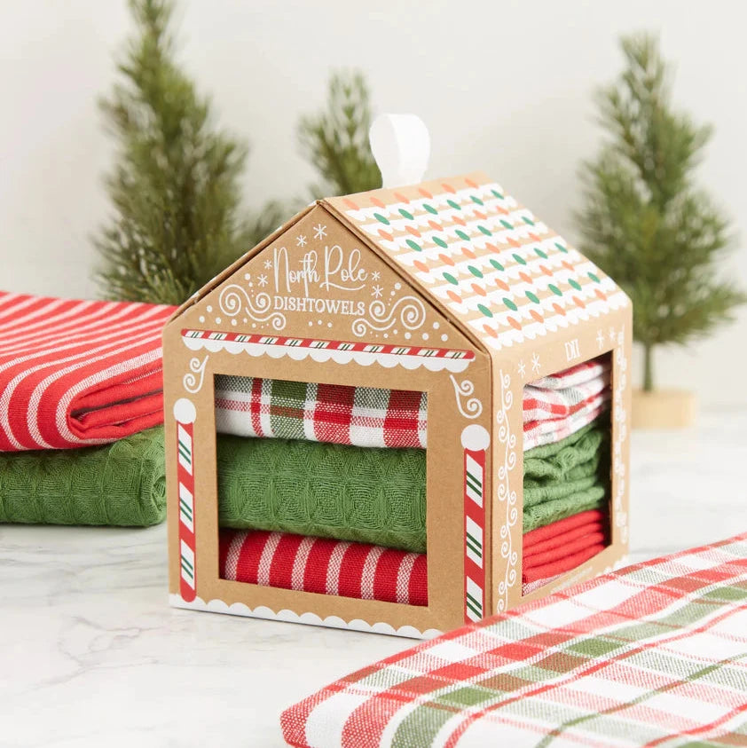 Packaged set of red, green, and white plaid towels in a house-shaped box with Christmas trees in the background.