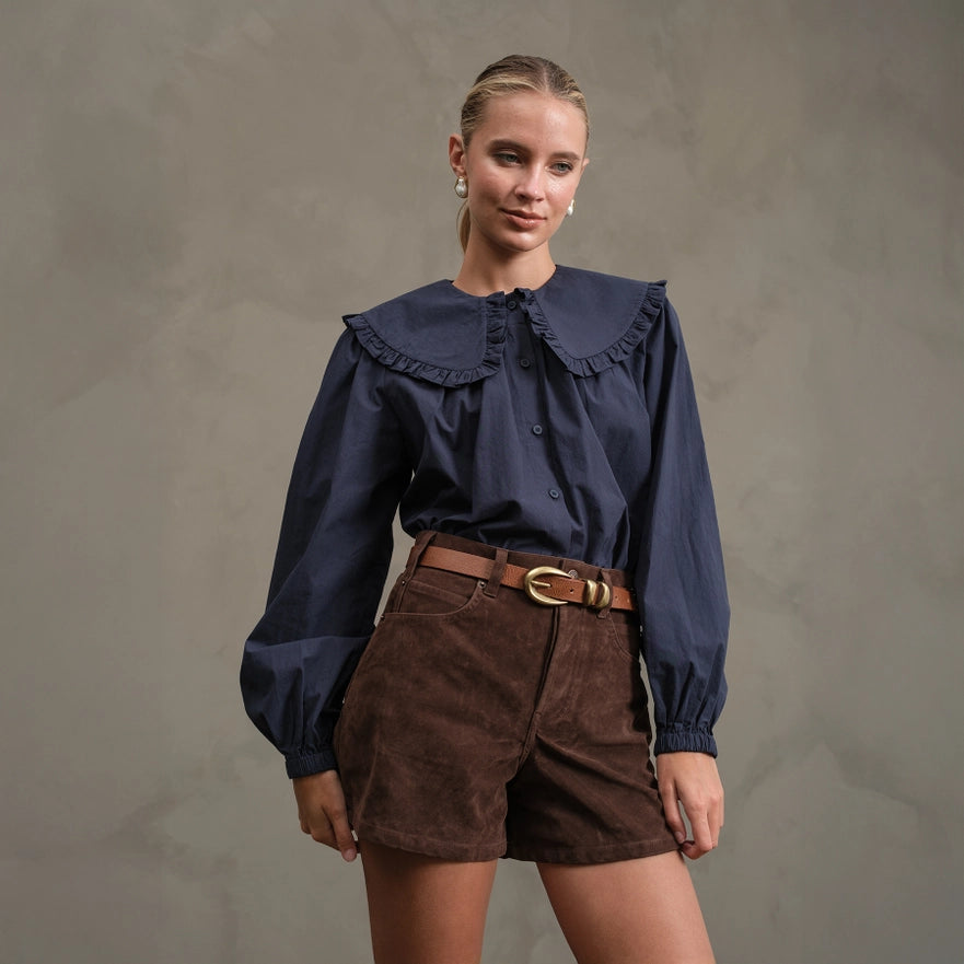 Woman wearing a navy blouse with ruffled collar and brown shorts against a gray background