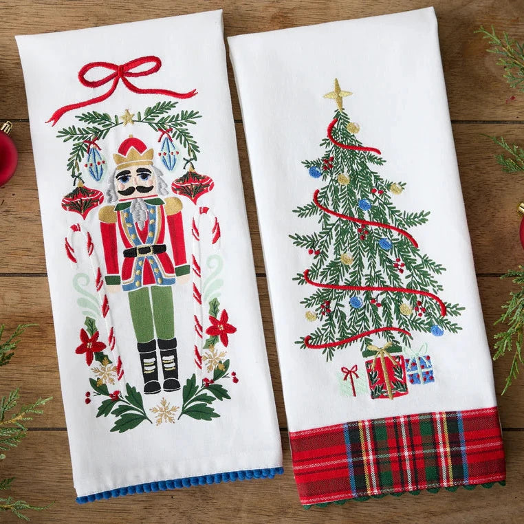 Two Christmas-themed kitchen towels with a nutcracker and Christmas tree design on a wooden surface.