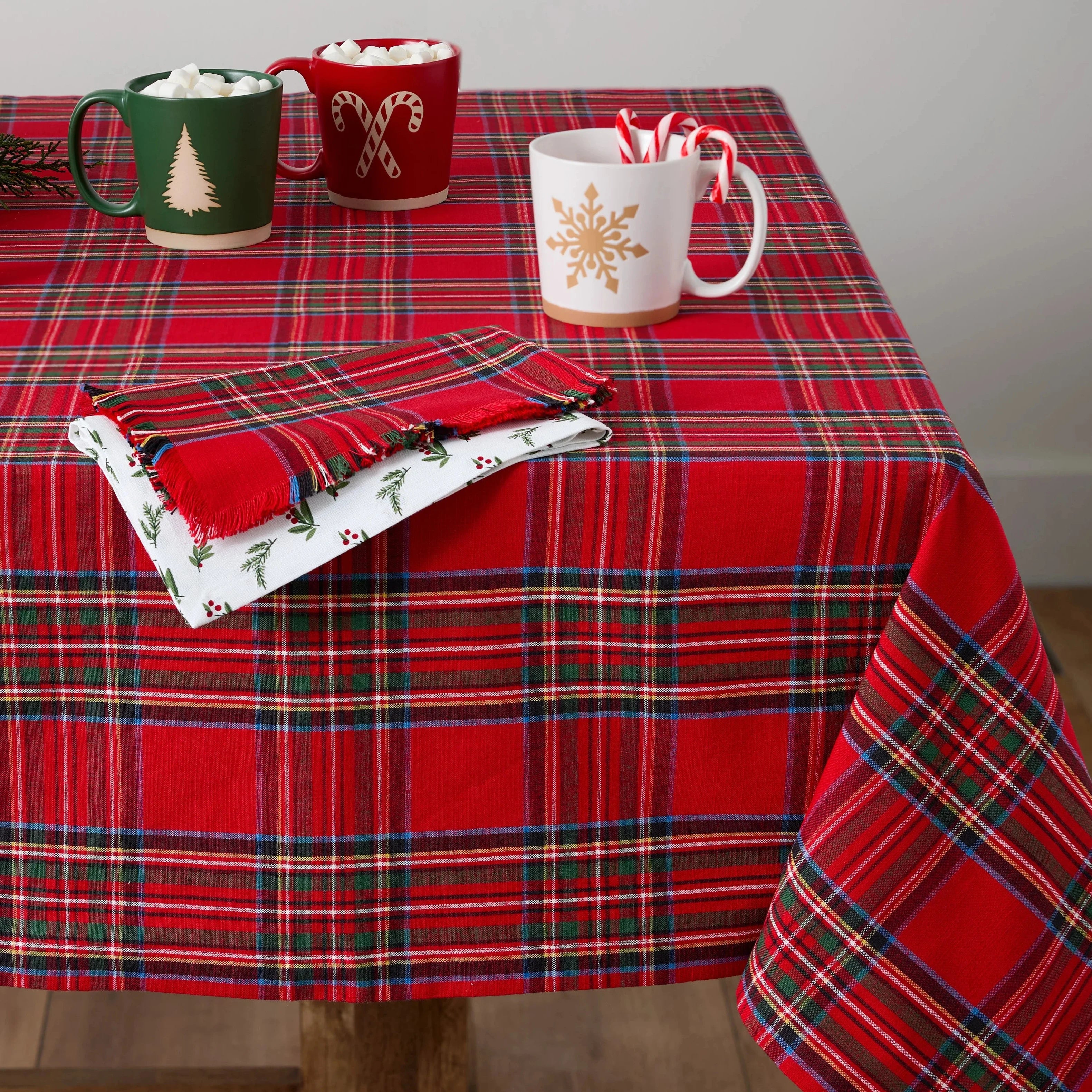 Red plaid tablecloth on a wooden table with Christmas mugs, cookies, and decorations.